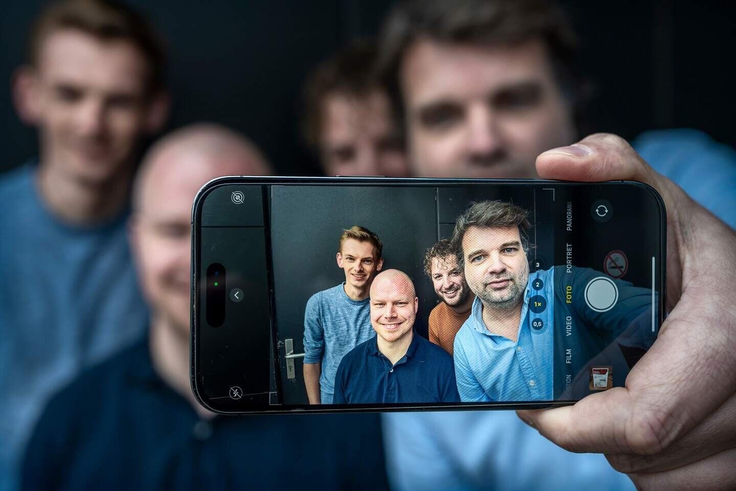 Four members of the app team taking a selfie, photographed through the phone's screen. Sebastiaan is at the back, with curly hair.